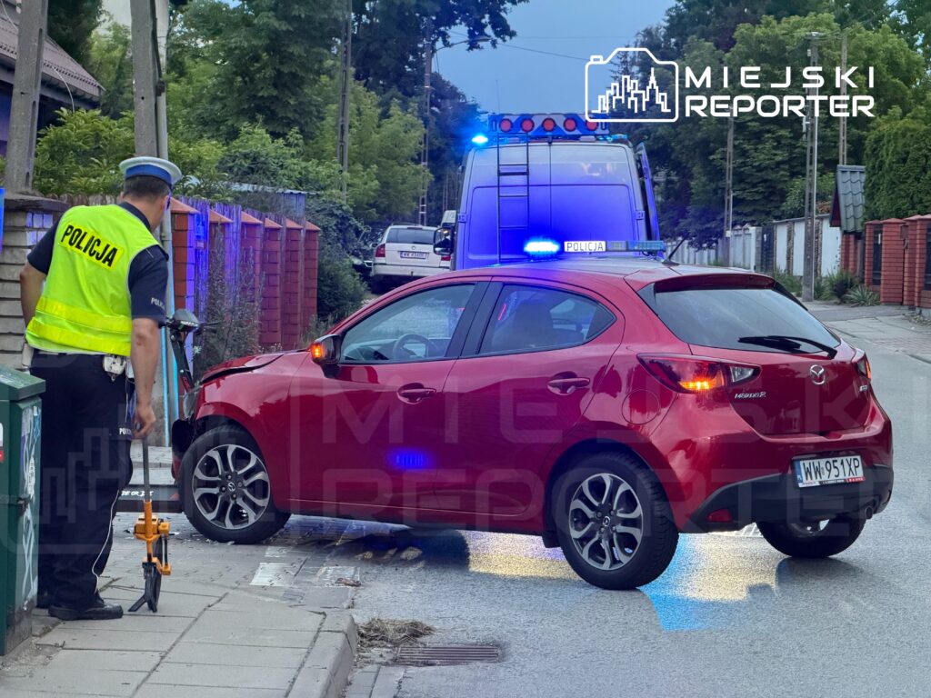 Policjant w kamizelce odblaskowej kontroluje uszkodzony czerwony samochód osobowy na ulicy, obok radiowozu z włączonymi sygnałami.