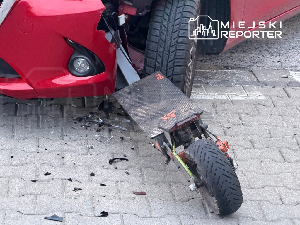 Czerwony samochód osobowy z uszkodzoną przednią częścią stoi na chodniku obok rozbitej hulajnogi.
