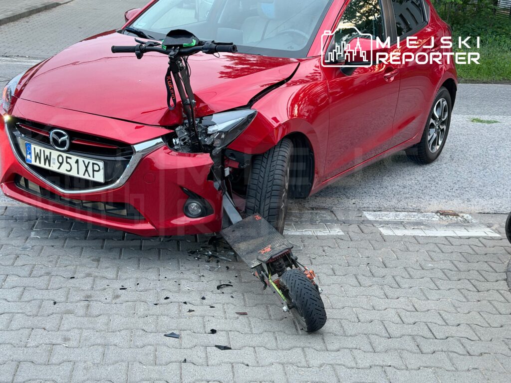 Czerwony samochód osobowy z uszkodzoną przednią częścią i odwróconą hulajnogą leżącą obok na chodniku.