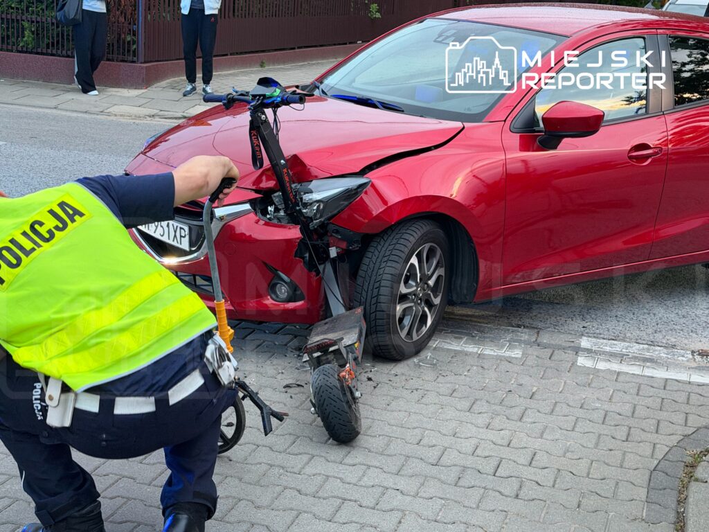 Policjant w kamizelce odblaskowej bada uszkodzone auto osobowe po kolizji z hulajnogą na chodniku.