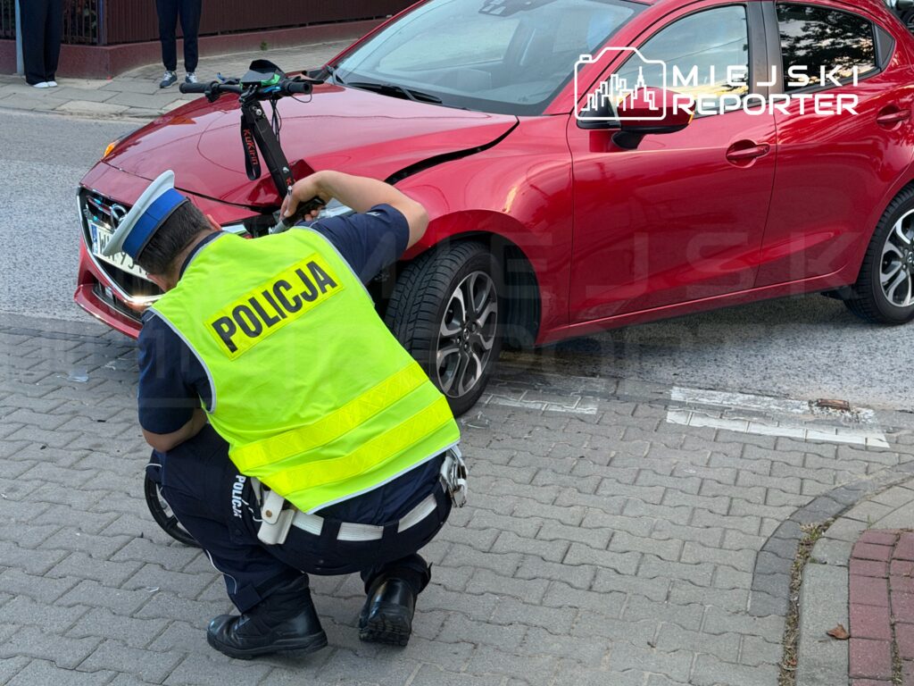 Policjant w kamizelce odblaskowej bada uszkodzone czoło czerwonego samochodu na chodniku.