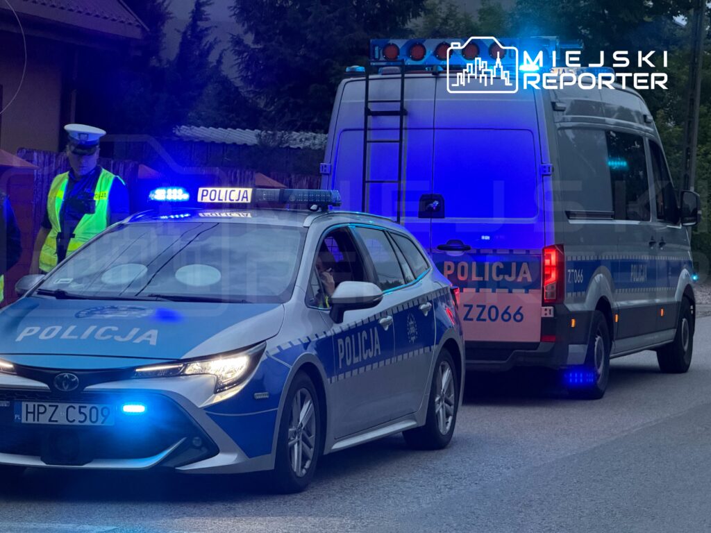Policjanci w odblaskowych kamizelkach kontrolują ruch obok radiowozu z włączonymi sygnałami świetlnymi.