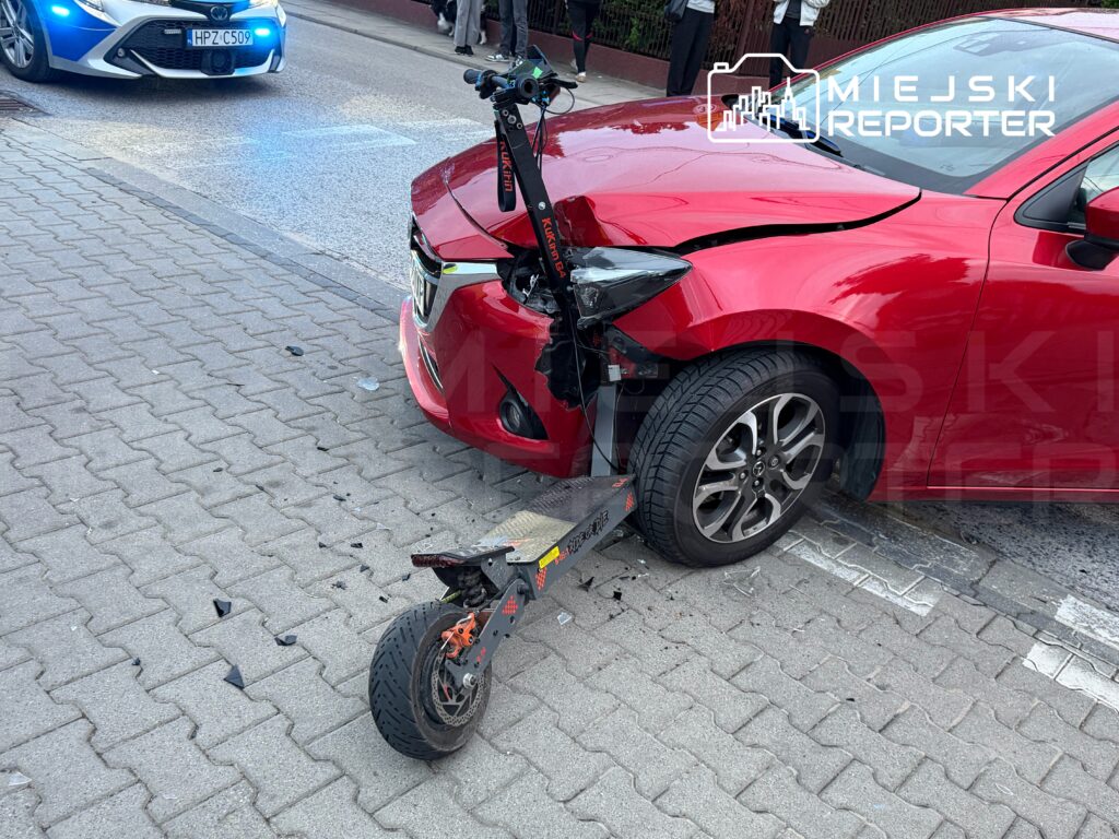 Czerwony samochód osobowy z uszkodzoną przednią częścią stoi na chodniku obok uszkodzonej hulajnogi elektrycznej.