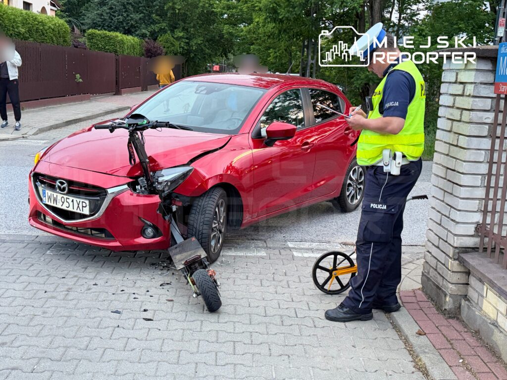 Policjant w kamizelce odblaskowej notuje zdarzenie obok uszkodzonego czerwonego samochodu osobowego.