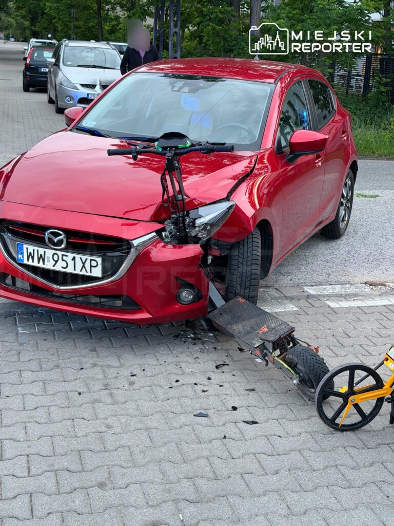 Czerwony samochód osobowy z uszkodzoną przednią częścią zaparkowany na chodniku obok uszkodzonego hulajnogi.