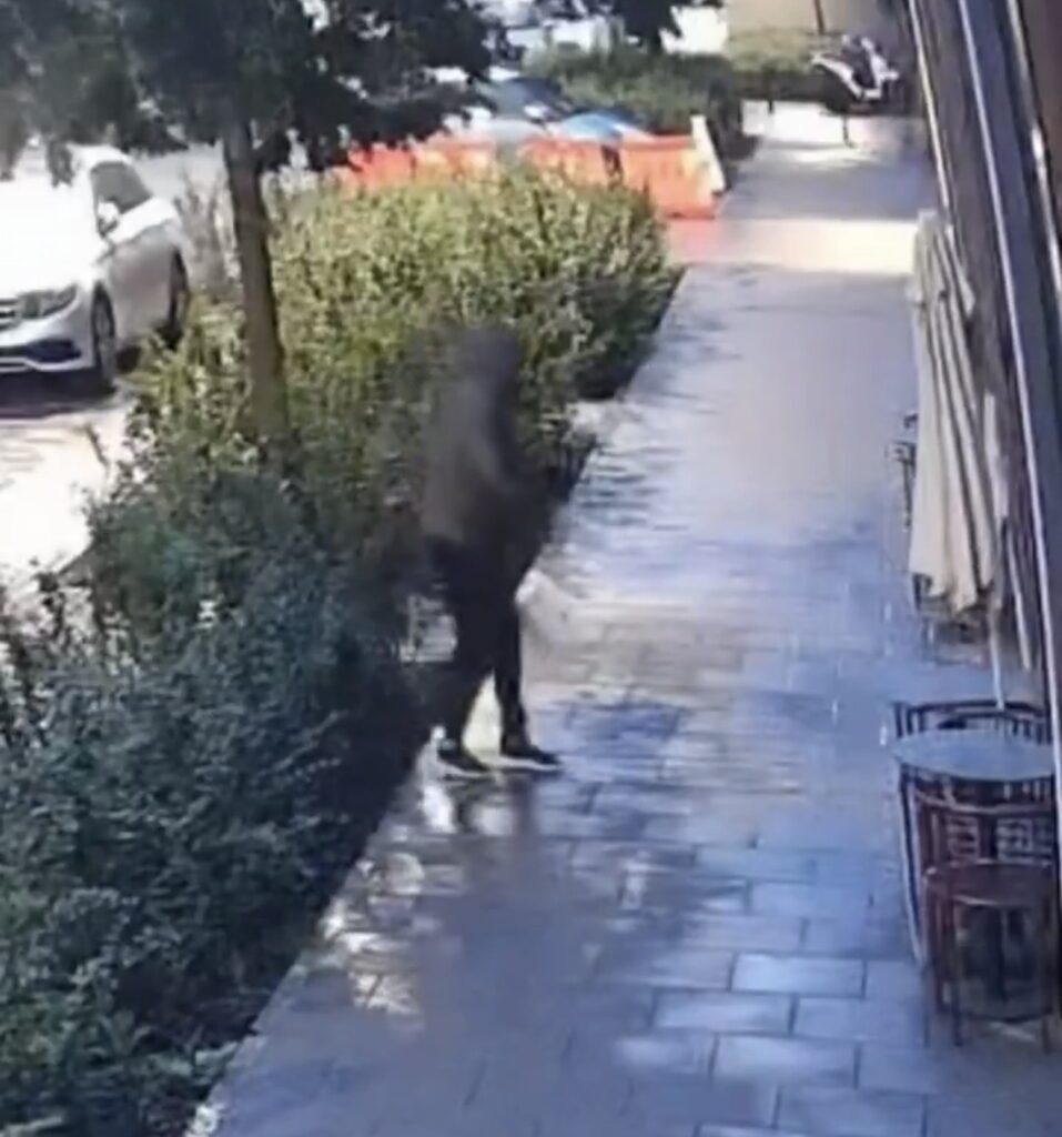 Mężczyzna w ciemnym ubraniu idący po mokrej ulicy obok zielonych krzewów, w tle samochody i barierki.