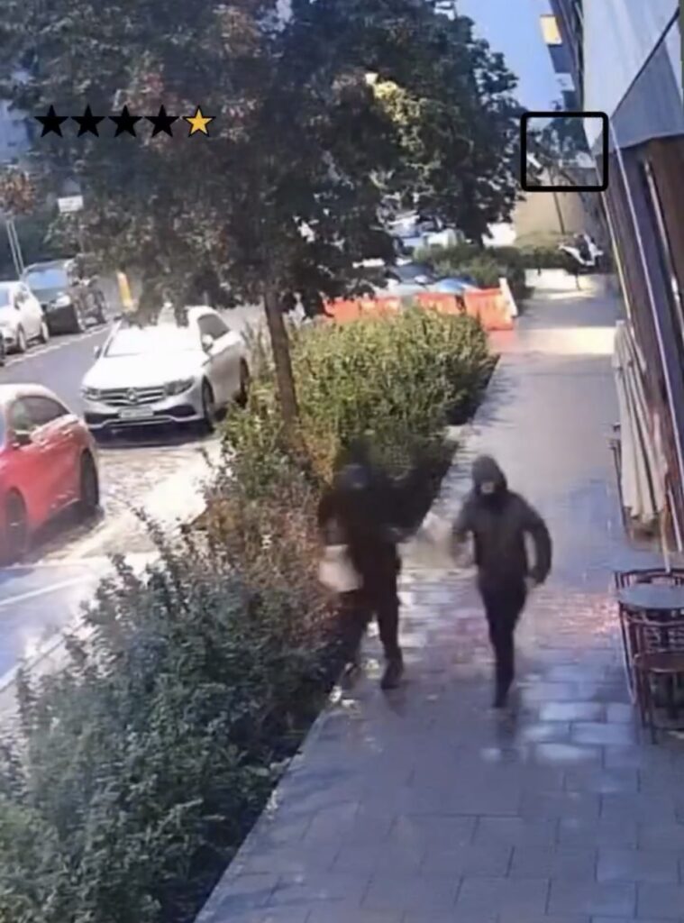 Dwaj mężczyźni w ciemnych ubraniach biegną chodnikiem, mijając zaparkowane samochody i zarośla.