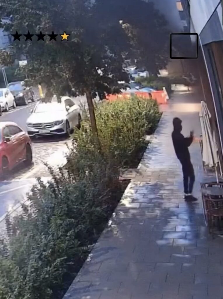 Mężczyzna w czarnej bluzie stoi na chodniku, trzymając telefon, w tle samochody i zieleń.