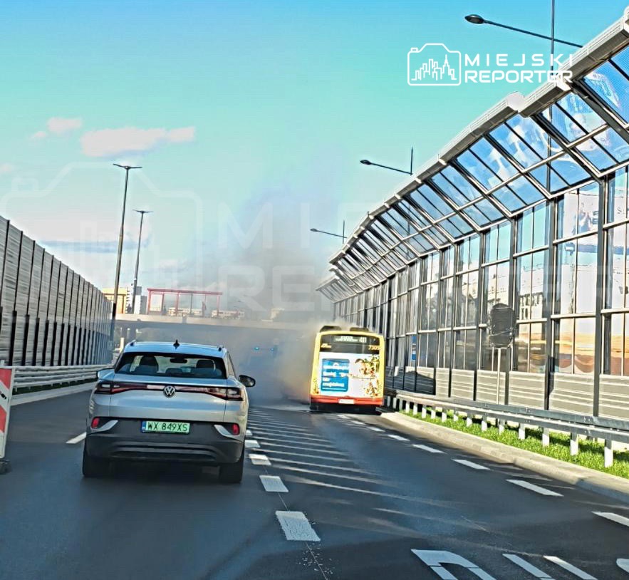 Samochód osobowy porusza się w kierunku autobusu, z którego wydobywa się dym, na ruchliwej drodze w mieście.