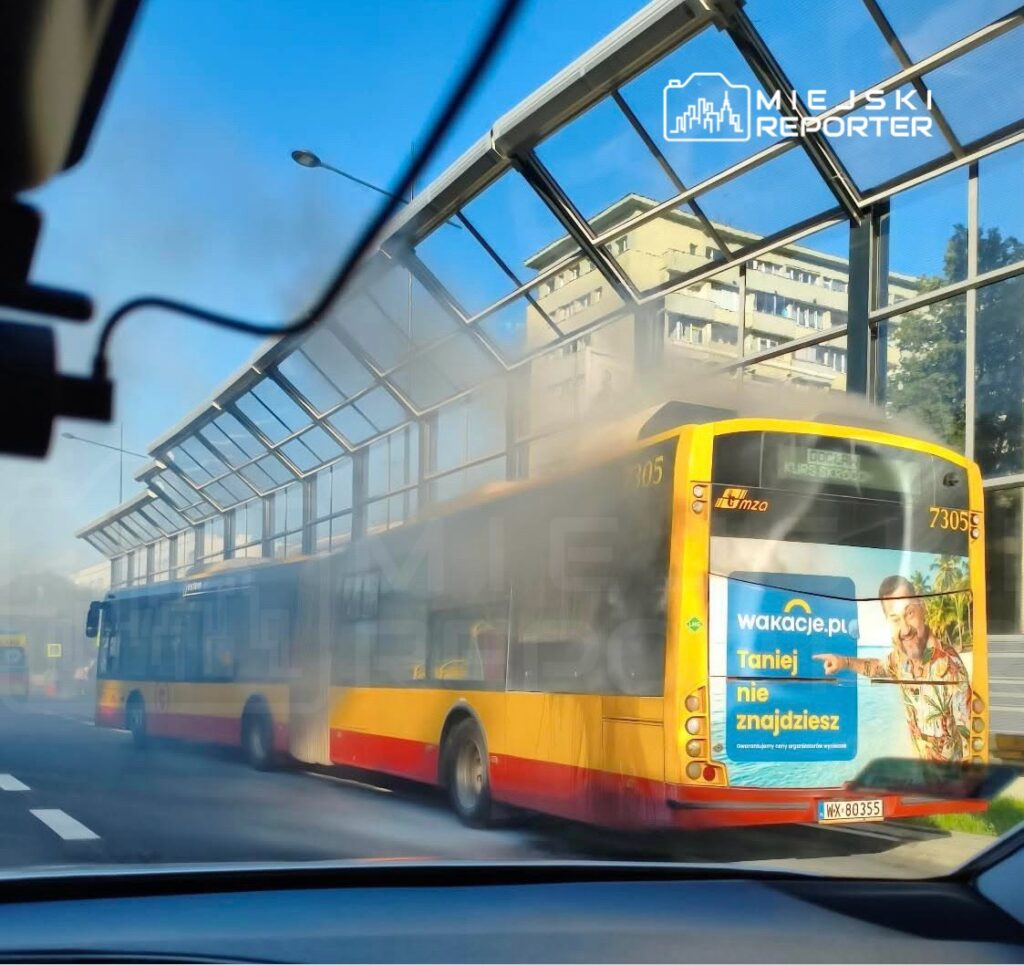 Autobus komunikacji miejskiej wydobywa dym na przystanku, obok przeszklonej wiaty.