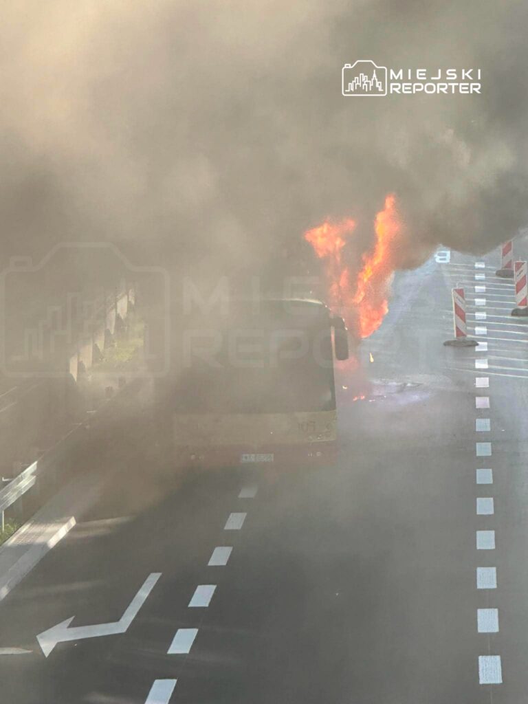 Autobus płonący na drodze, otoczony gęstym dymem, w tle widoczne są znaki drogowe.