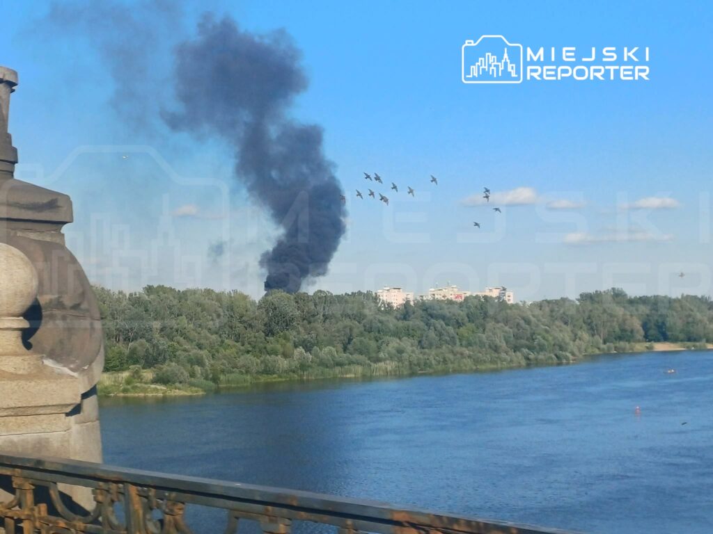 Czarny dym unoszący się nad drzewami wzdłuż rzeki, w tle widoczne ptaki i zabudowania.
