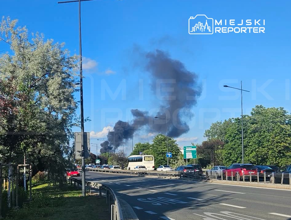 Czarny dym unoszący się nad miastem w pobliżu ruchliwej drogi, otoczony zielenią drzew.