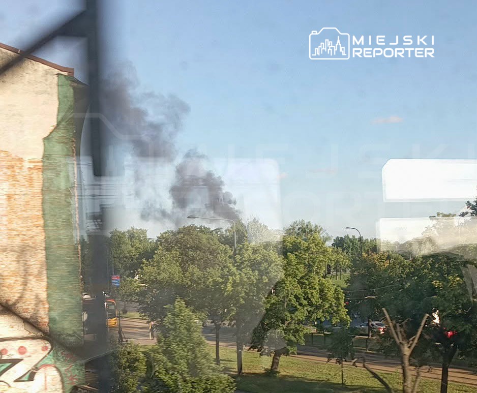 Czarny dym unoszący się nad drzewami w miejskim otoczeniu, widoczny z okna budynku.