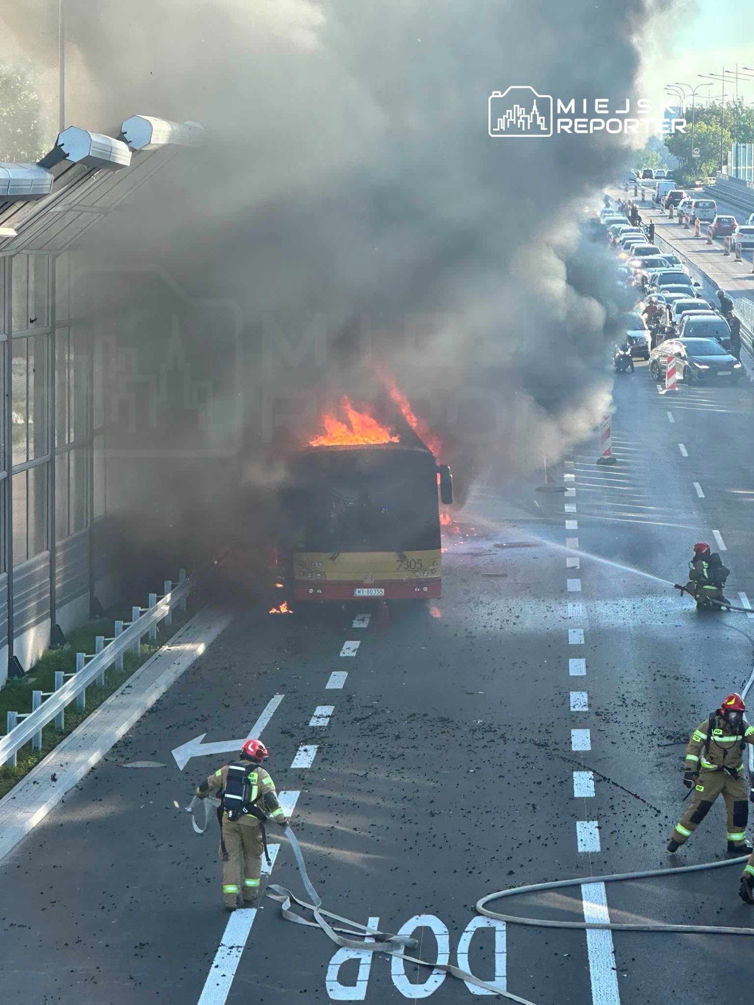Pożar autobusu na jezdni, strażacy gaszą płomienie, w tle widoczne zablokowane samochody.