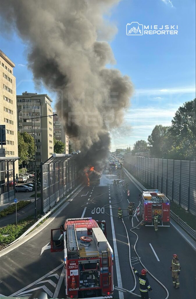 Pożar pojazdu na drodze, w tle strażacy gaszą ogień, a dym unosi się w powietrzu.