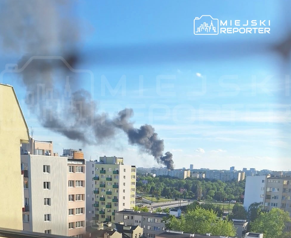 Gęsty, czarny dym unoszący się z obszaru zabudowań miejskich, w tle widać zieloną przestrzeń i budynki.