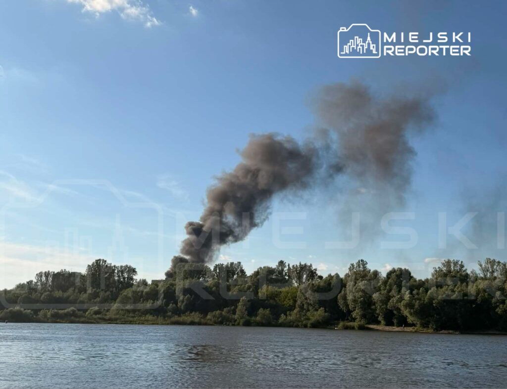 Gęsty czarny dym unosi się z zielonego wzgórza nad rzeką w słoneczny dzień.