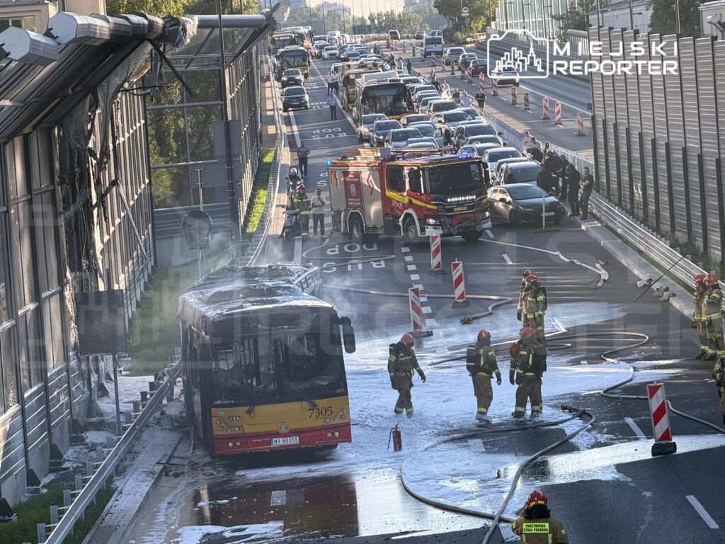 Spalony autobus na jezdni, strażacy gaszą pożar, a w tle zablokowany ruch drogowy.