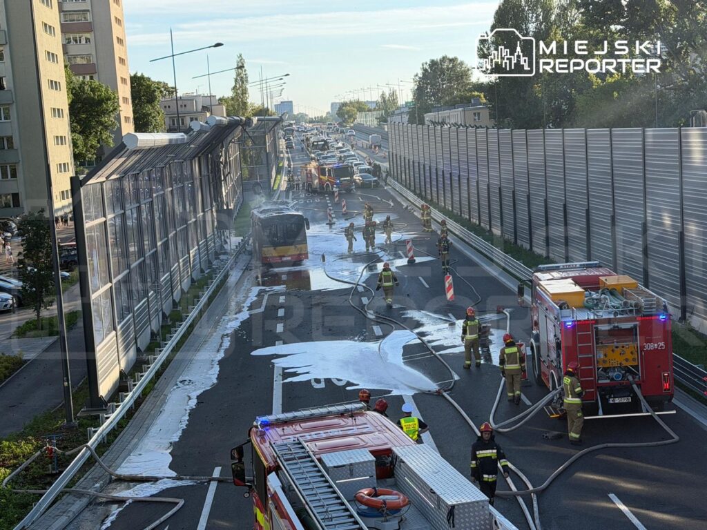 Strażacy gaszą pożar autobusu na drodze, obok widoczny wąż strażacki i rozlane środki gaśnicze.