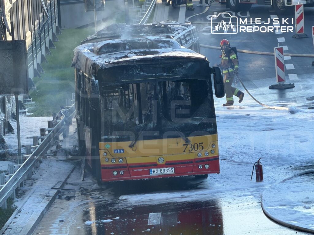 Spalony autobus z widocznymi śladami pożaru, obok strażak gaszący ogień na jezdni.