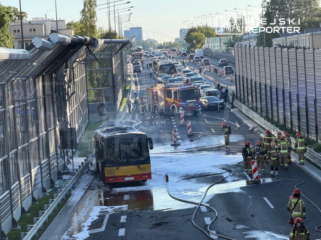 Spalony autobus stoi przy drodze, obok pracują strażacy, a w tle widać zablokowany ruch samochodowy.