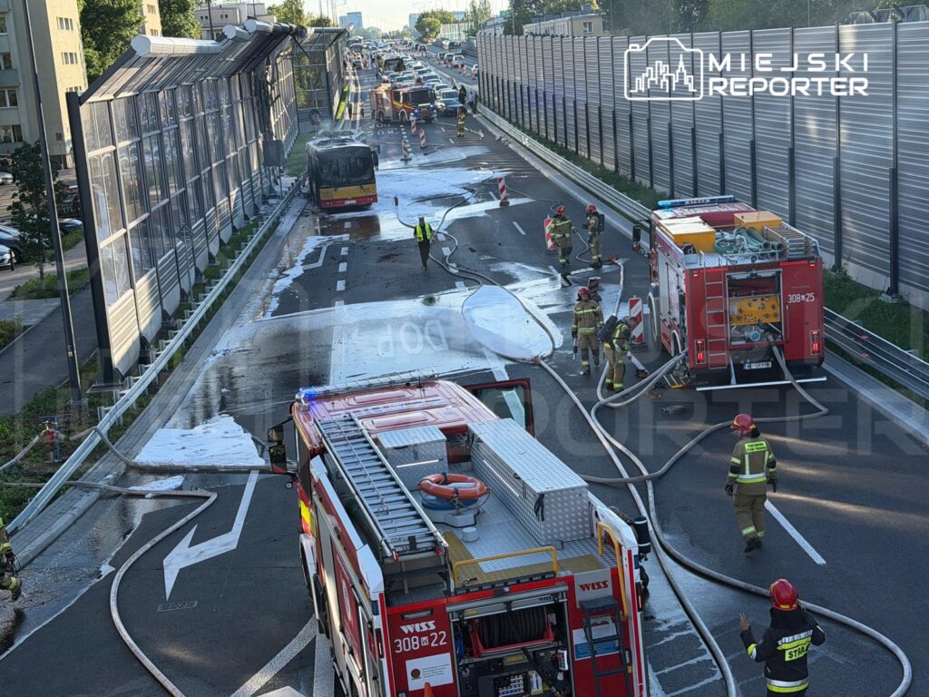 Grupa strażaków pracuje nad gaszeniem pożaru autobusu na ruchliwej drodze, obok stoją dwa wozu strażackie.
