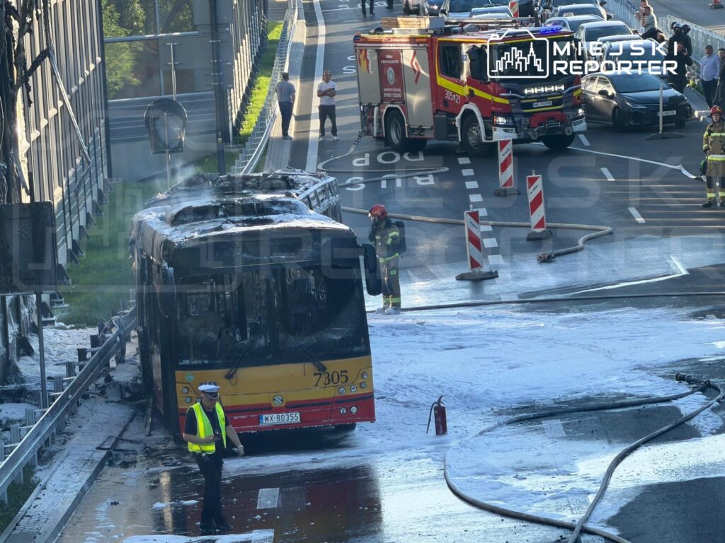 Zniszczony autobus przystankowy, obok którego pracują strażacy, a w tle widać inne pojazdy na drodze.