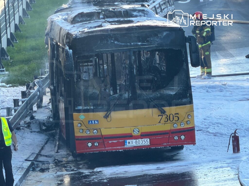 Spalony autobus z numerem taborowym 7305 stoi na poboczu drogi, obok strażaków gaszących pożar pianą gaśniczą.