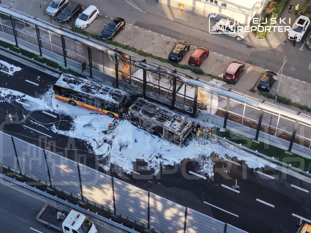 Spalony autobus komunikacji miejskiej jest gaszony przez strażaków na jezdni, otoczony przez płyn gaśniczy.