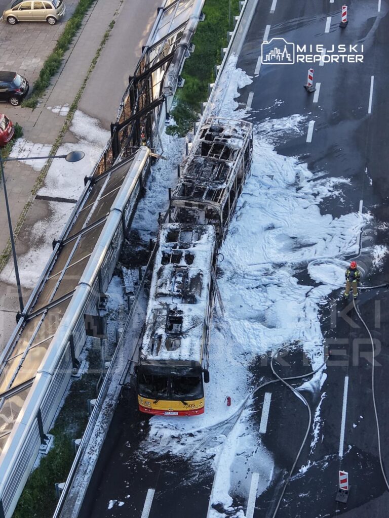 Spalony autobus leży na jezdni, pokryty pianą gaśniczą, obok strażak z wężem. W tle widoczna przeszklona konstrukcja.