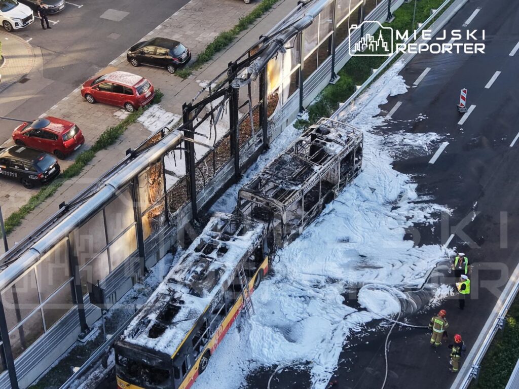 Spalony autobus leży na jezdni, otoczony przez strażaków używających piany gaśniczej oraz uszkodzone ogrodzenie.