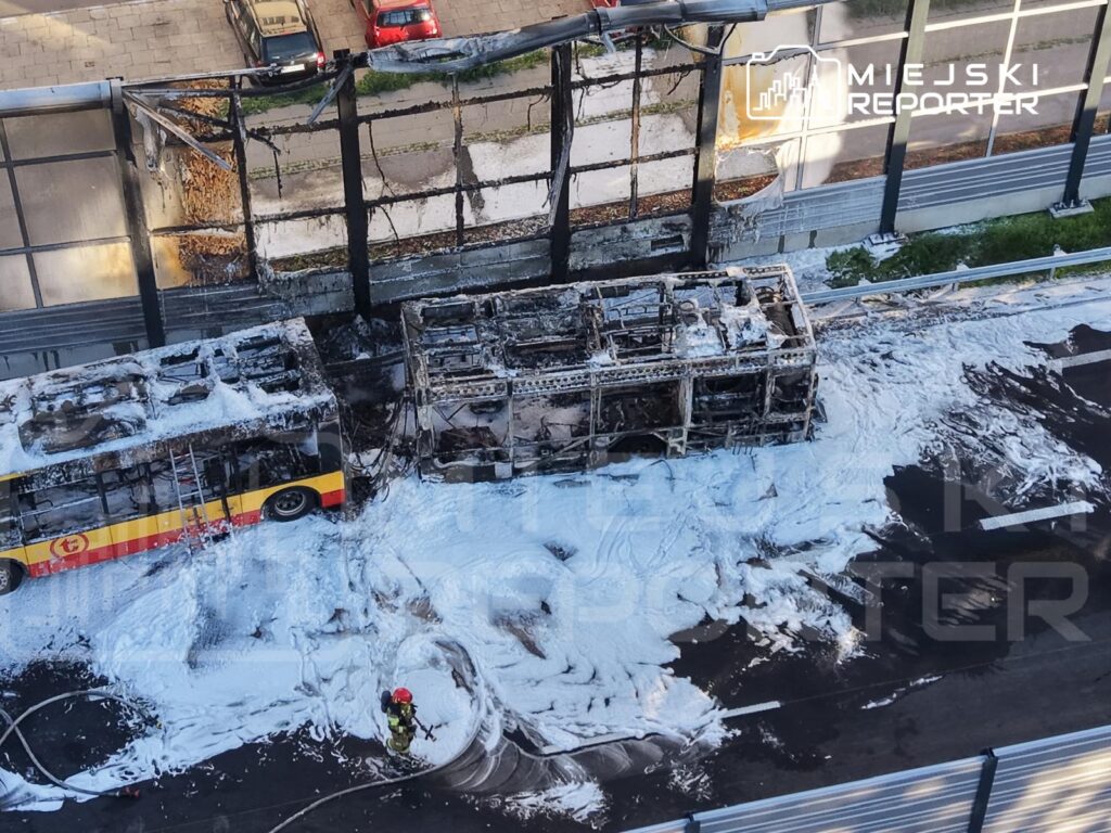 Spalony autobus wypełniony pianą gaśniczą, obok strażak w hełmie pracujący nad ugaszeniem pożaru.