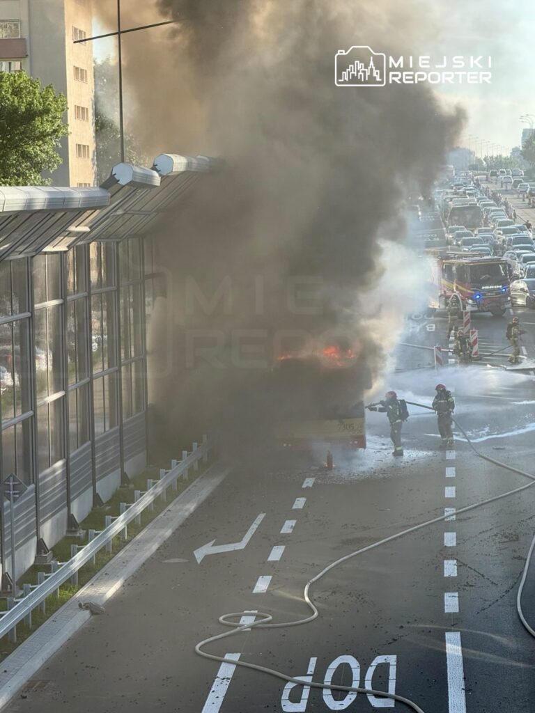 Strażacy gaszą ogień w płonącym autobusie na ruchliwej drodze, w tle widoczne zablokowane pojazdy.