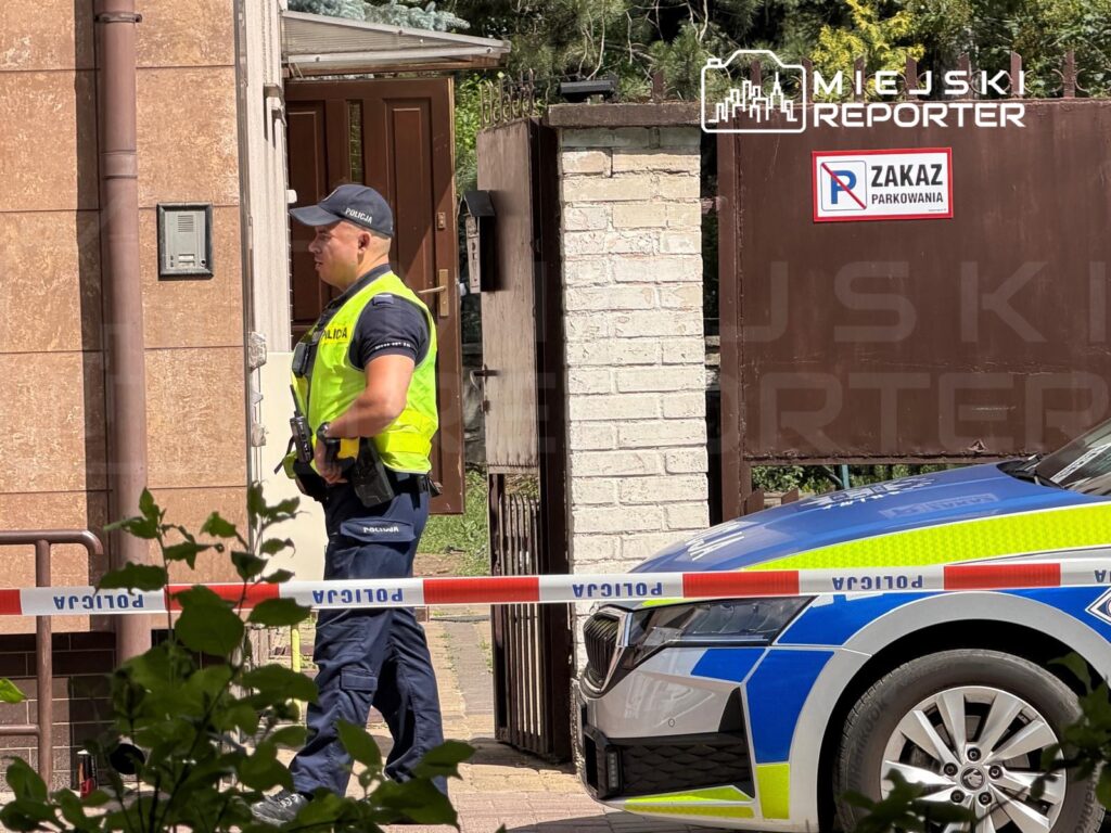 Policjant w kamizelce odblaskowej stoi przy bramie, obok oznakowanego radiowozu, w otoczeniu zieleni.