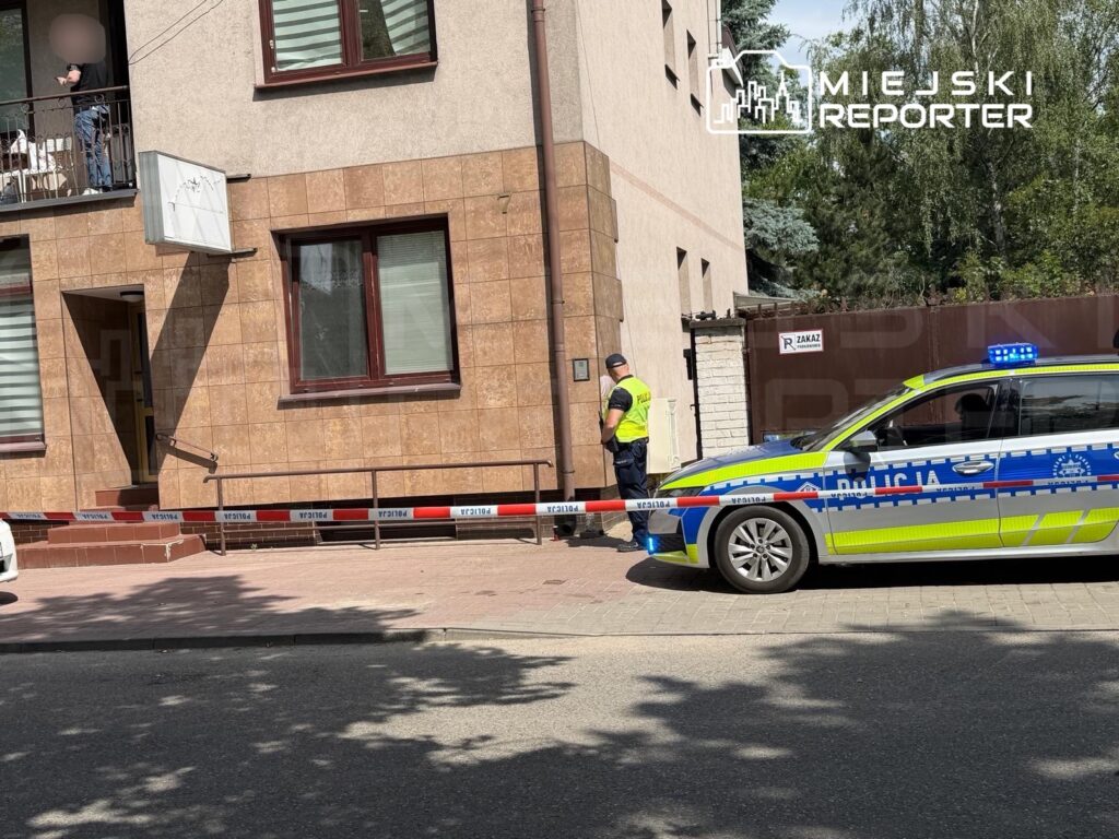 Policjant zabezpiecza miejsce zdarzenia przy budynku mieszkalnym, obok stoi radiowóz policyjny z włączonymi sygnałami.