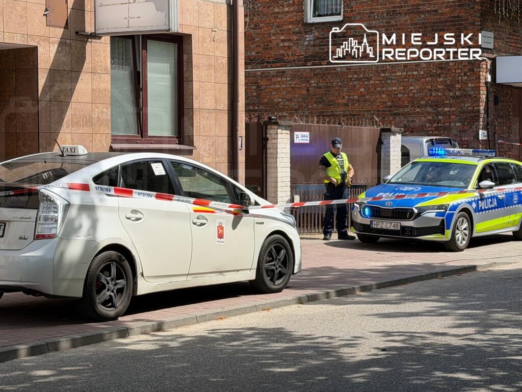 Na chodniku stoi białe taxi z taśmą policyjną, obok policjant w kamizelce odblaskowej rozmawia z kolegą przy radiowozie.