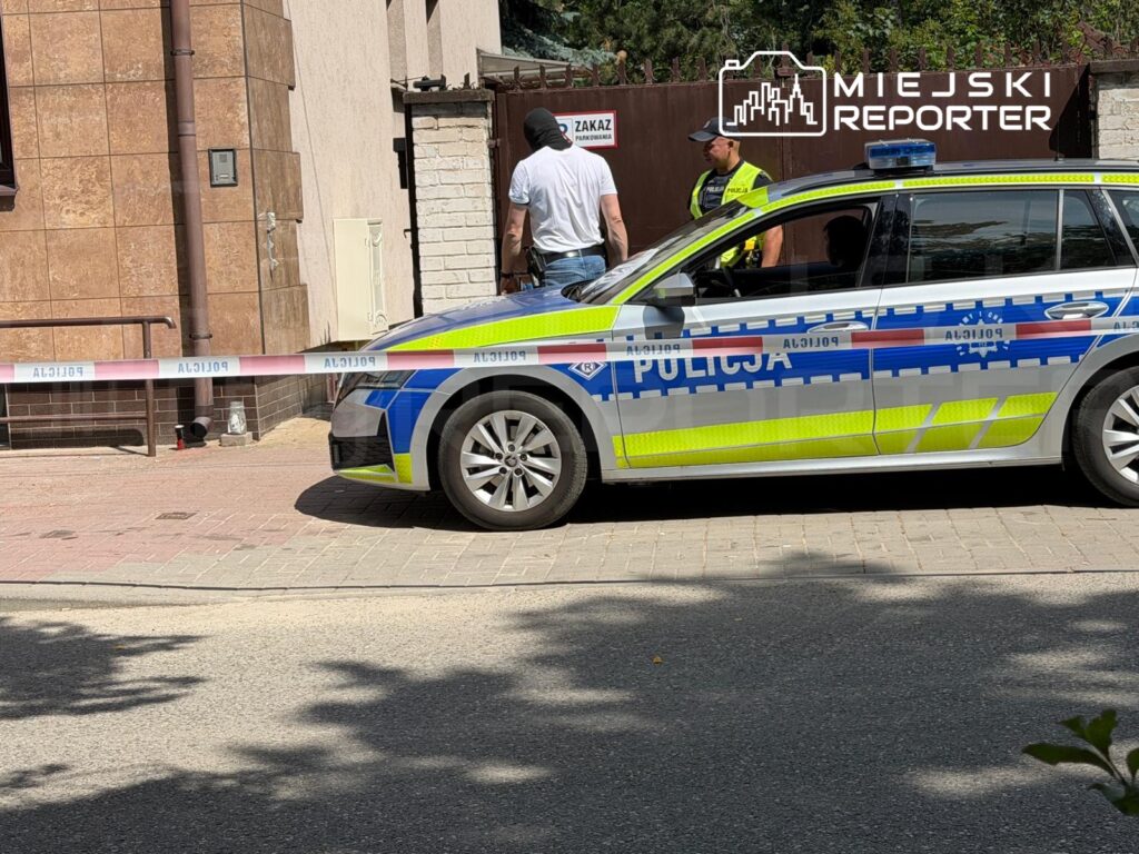 Mężczyzna w białej koszulce rozmawia z policjantem obok radiowozu przy zabezpieczonym miejscu z taśmą.
