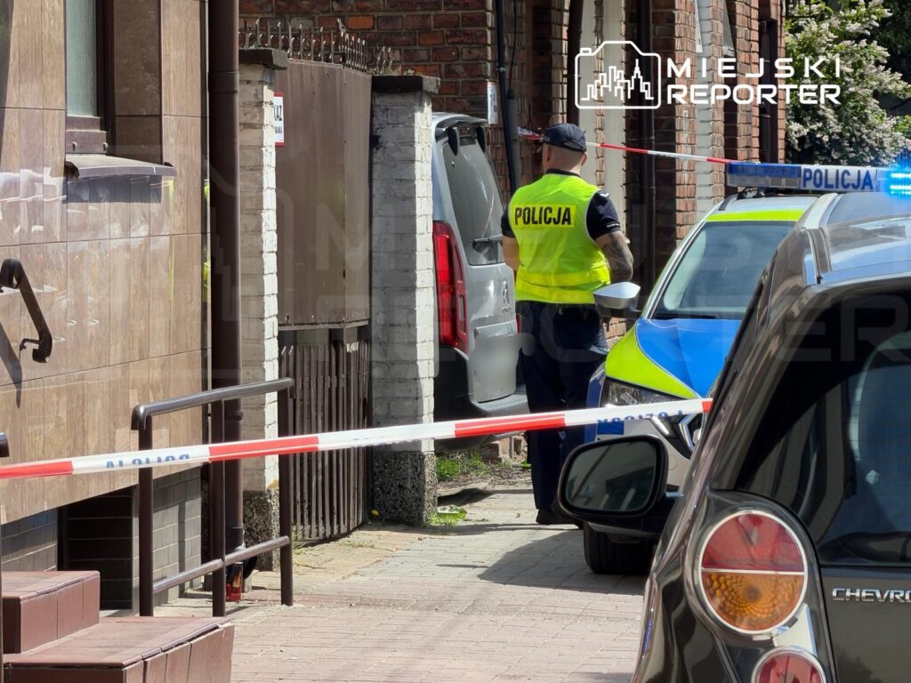Policjant w kamizelce odblaskowej zabezpiecza miejsce zdarzenia, obok zaparkowane samochody i taśmy policyjne.