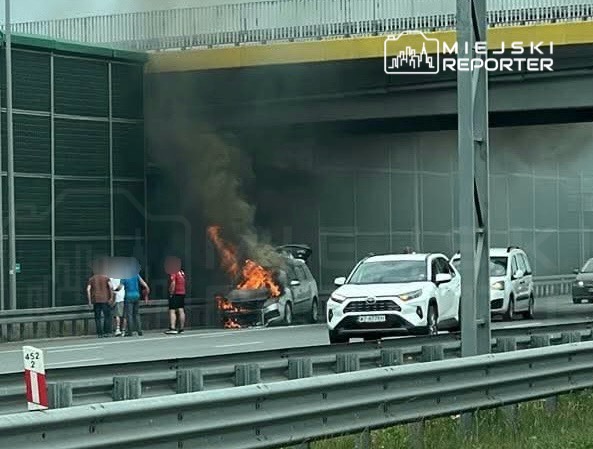 Samochód płonący na pasie ruchu, obok grupka osób obserwujących zdarzenie, w tle inne pojazdy.