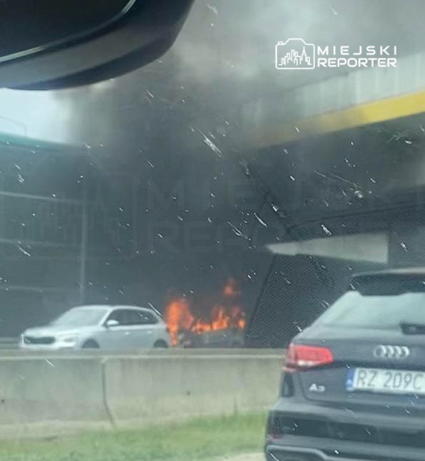 Pojazd płonący pod wiaduktem, unoszący się czarny dym, w tle stojące samochody na drodze.