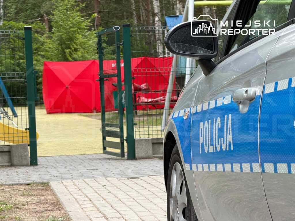 Policyjny samochód zaparkowany przy placu zabaw z czerwonymi namiotami w tle, otoczony zielenią drzew.