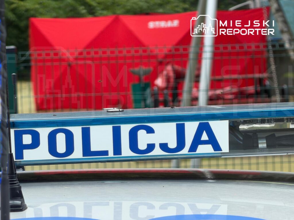 Policjant stojący przy radiowozie z napisem "POLICJA", w tle czerwony namiot.