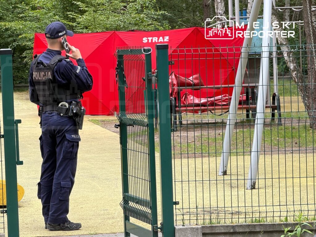 Policjant w mundurze rozmawia przez telefon przy ogrodzeniu placu zabaw z czerwonym namiotem w tle.
