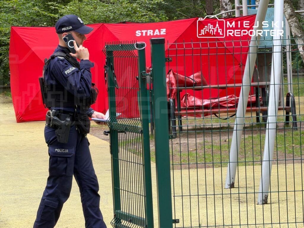 Policjant w mundurze rozmawia przez telefon obok zamkniętej bramy w parku, w tle czerwony namiot.