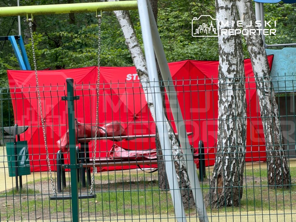Zielony plac zabaw z huśtawkami, w tle czerwony namiot oraz zadaszone urządzenia do zabawy, otoczone drzewami.