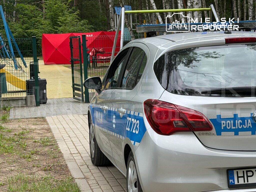 Policyjny samochód zaparkowany przy placu zabaw z huśtawkami i zjeżdżalnią w otoczeniu drzew.