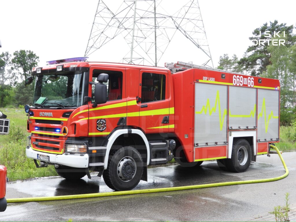 Czerwony wóz strażacki marki Scania z numerem 669M66 stoi na mokrej nawierzchni, otoczony zielonymi drzewami.