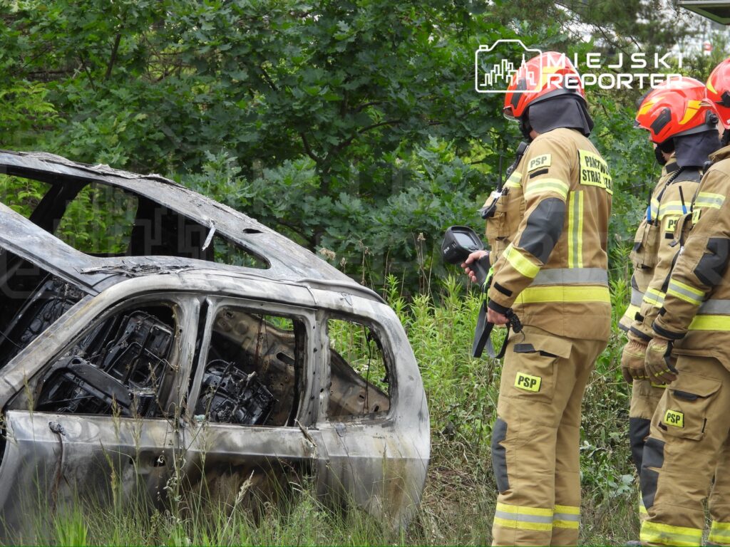 Strażacy w mundurach ochronnych badają spalone auto w zaroślach, używając sprzętu pomiarowego.