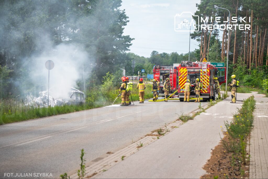 Grupa strażaków gasi pożar wraku samochodu na poboczu drogi, obok stojące wozy strażackie.